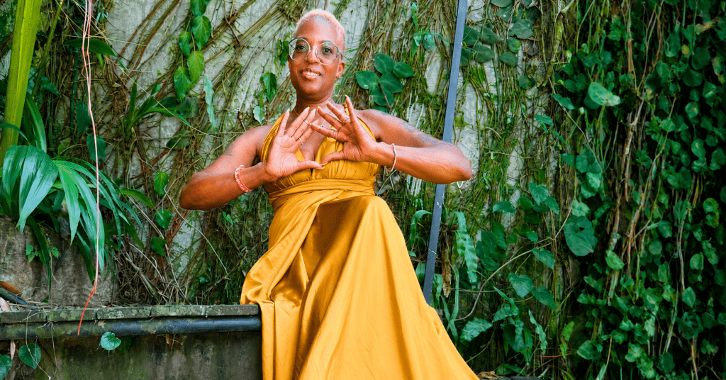 A woman in a flowing golden dress smiles while seated among lush tropical greenery, celebrating personal growth, resilience, and a life intentionally lived through travel and self-discovery.