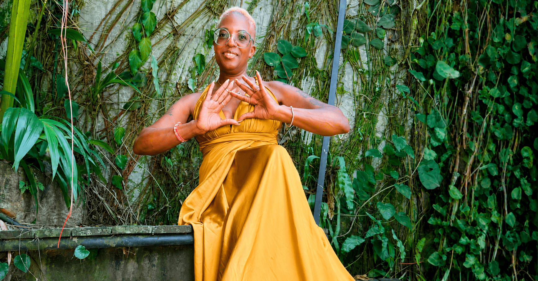 A woman in a flowing golden dress smiles while seated among lush tropical greenery, celebrating personal growth, resilience, and a life intentionally lived through travel and self-discovery.